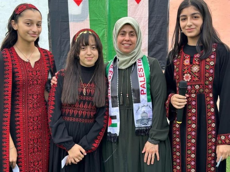 Three students in traditional embroidered dresses stand with a teacher wearing a Palestine scarf