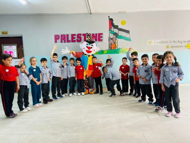 Students stand in a semicircle with a costumed character under a “Palestine” wall decoration